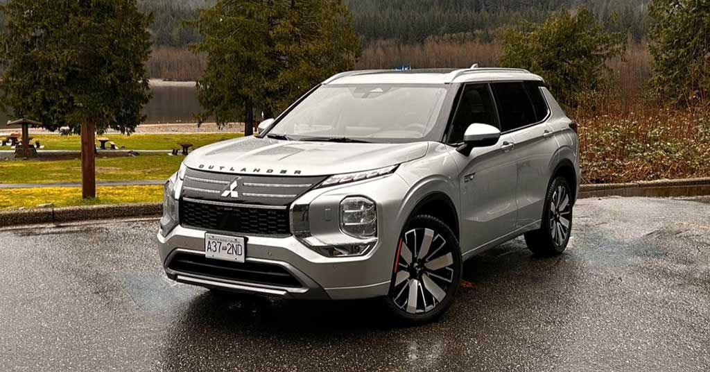 2026-Outlander-PHEV - Sask Valley Voice Front-three-quarter view of a silver Mitsubishi Outlander SUV parked on a wet pavement with trees and a lake in the background, daytime outdoor scene.